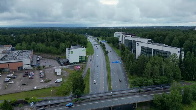 Cinematic aerial drone footage over Pohjois-Haaga, Helsinki, Finland, featuring the Veikkaus building, surrounded by lush green summer landscapes under a cloudy sky.