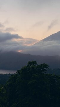 Vertical video. Mount Abang And Lake Batur Beneath Flowing Clouds at Early Sunrise, Soft Morning Light Illuminates Volcanic Highlands And Lush Forested Terrain