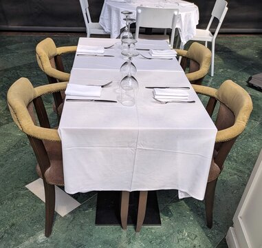 Mesa rectangular con mantel blanco y sillas acolchadas con patas de madera de dise&ntilde;o cl&aacute;sico, en un restaurante o sal&oacute;n de hotel, con cubiertos, servilletas y copas de vino sobre la mesa.	