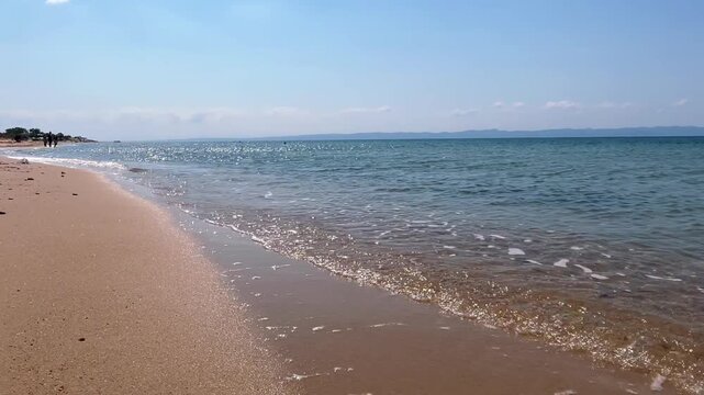 Low angle static shot of gentle sea waves with white foam washing onto a sandy beach under a bright blue sky in Saros Gulf, Edirne, Turkey.