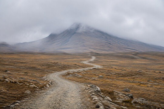 Winding gravel path on high plateau leading to misty mountain, vast landscape.