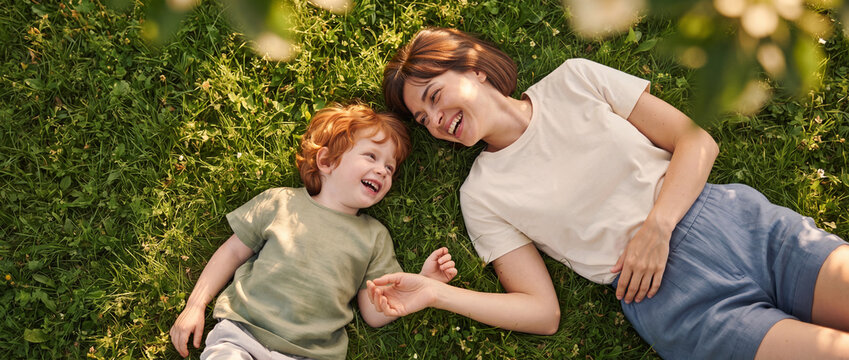 Mother and son laughing on green grass during a sunny summer day in the park, top view of happy smiling redhead toddler boy and cheerful mom relaxing outdoors together