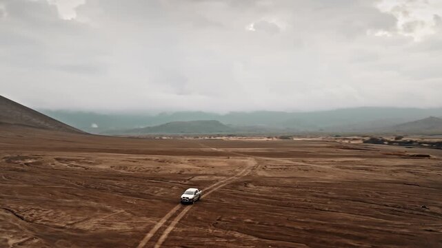 White truck driving on dirt track across the vast, rugged volcanic ash plain with Mount Yasur volcano slope looming under a cloudy sky in Tanna Island, Port Resolution. Aerial view drone panorama
