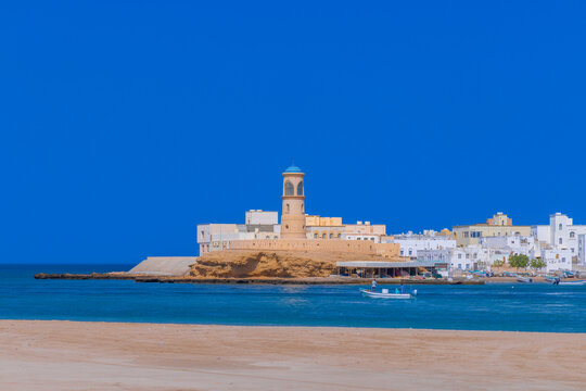 Sur Lighthouse stands at the shoreline with boats nearby in a clear sky over the coast of Oman