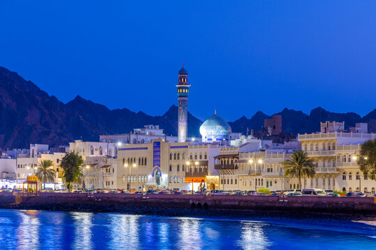 Evening view of Mutrah Corniche with buildings and mountains in the background and street lights illuminating the waterfront area