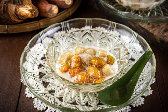 Homemade Thai Bua Loi Kaew dessert. Translucent orange sweet potato and taro tapioca balls in warm coconut milk soup. Served in glass bowls on a rustic wooden table with raw tubers. 