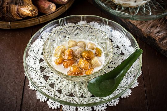 Homemade Thai Bua Loi Kaew dessert. Translucent orange sweet potato and taro tapioca balls in warm coconut milk soup. Served in glass bowls on a rustic wooden table with raw tubers. 