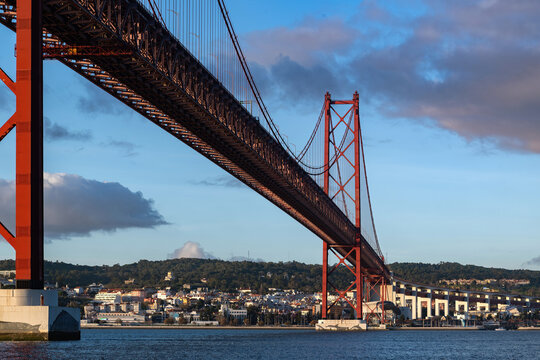 View of 25 April Bridge from low perspective along Tagus River with city outline in background during sunset light