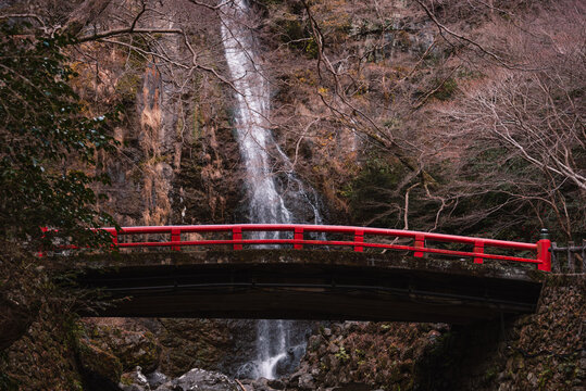 Red bridge spans rocky cliffs under Minoh Waterfall in winter with bare trees in Minoh Park, Japan