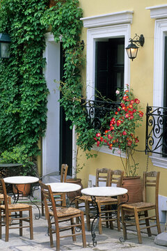 Chairs and tables set for visitors at a cafe terrace in Nafplio, with vibrant flowers against a yellow facade