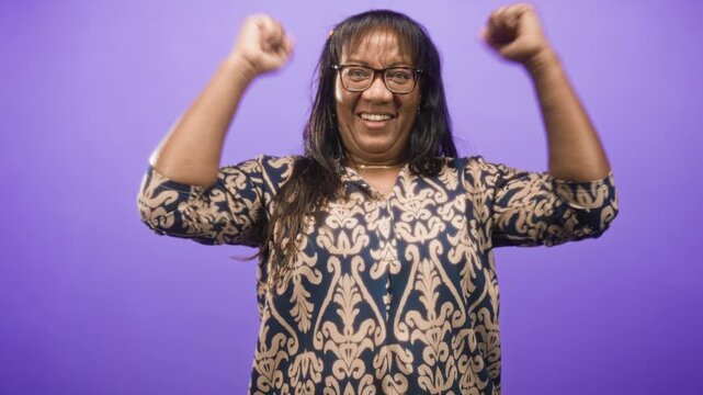 Woman middle age african american raising fists and hands to head, smiling in patterned blouse and glasses in purple studio; celebration joy.