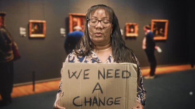 Woman holding cardboard sign reading we need a change, lips pursed in a museum building gallery with framed paintings visible; protest fatigue.