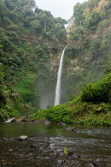 Tall jungle waterfall cascades over volcanic rock cliffs into crystal stream nature vista. © wahyu
