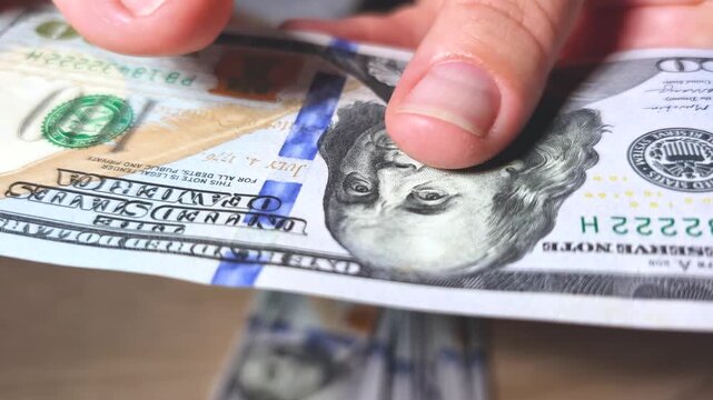 Close-up video of female hands taking a hundred dollar bill from a stack and carefully examining its texture and authenticity. Financial, banking, cash, payment, and counterfeit detection concept.