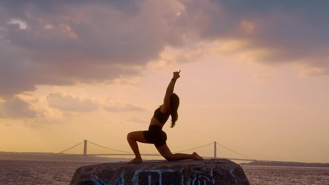 Woman practicing crescent lunge yoga pose at sunset in New York. Silhouette of person on concrete pier against Verrazzano Narrows Bridge and golden sky.