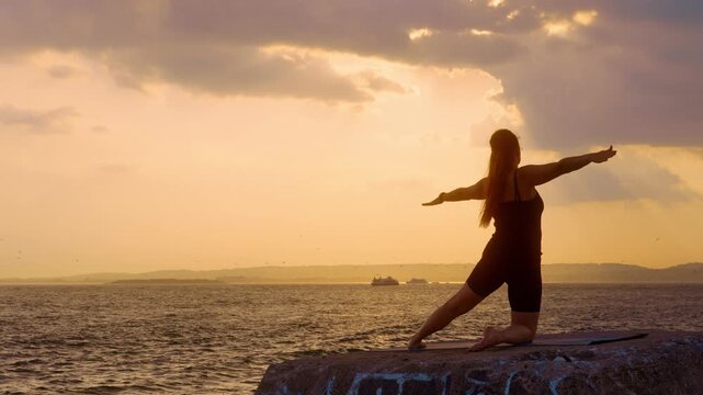 Woman practicing kneeling yoga stretch facing the water. Female silhouette extending arms sideways on stone ledge, golden sun rays through clouds, sea backdrop.