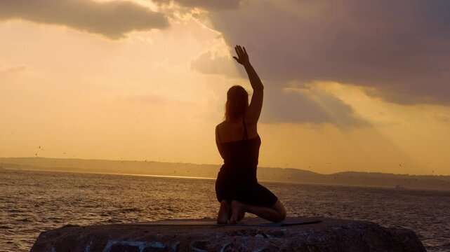Woman reaching arm up in kneeling yoga pose outdoors. Back view silhouette raising single hand, sunbeams breaking through cloudy sky, coastal landscape.