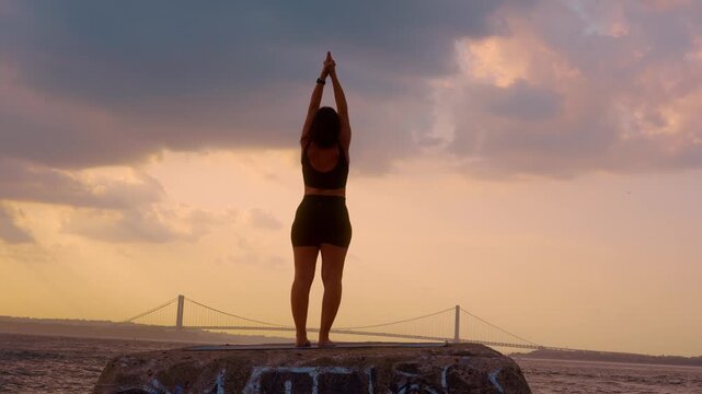 Woman doing standing yoga pose with raised hands at sunset in New York. Back view of female figure on stone block near water with Verrazzano Narrows Bridge in distance.