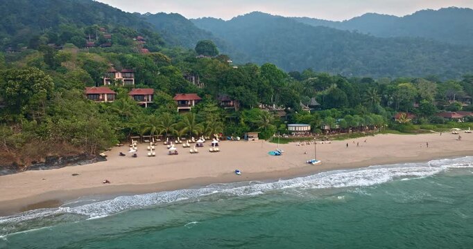 Koh Lanta beautiful sandy beach fringed by lush green jungle covered hills featuring a luxury resort with bungalows and palm trees, calm ocean waves. Summer vacation in Thailand. Drone shot panorama