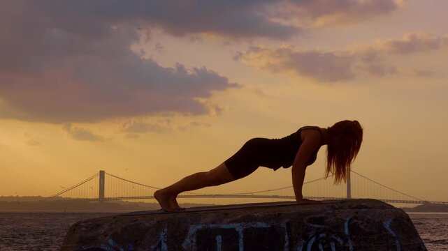 Woman holding plank position on stone pier near New York bridge. Full length side view silhouette of female doing yoga pushup exercise by sea facing Verrazzano landmark.