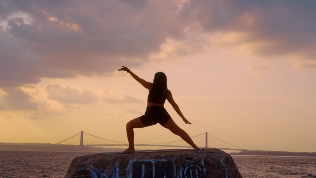 Woman in warrior yoga pose during sunset along New York bay. Silhouette performing side posture on pier with Verrazzano Narrows Bridge in cloudy background.