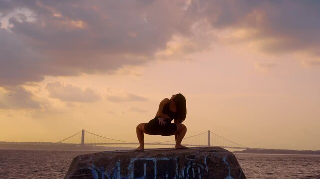 Woman executing deep squat yoga pose at sunset in New York. Wide stance posture on stone edge with dark sky and Verrazzano Narrows Bridge background.
