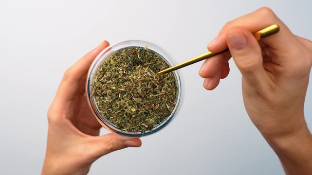 Gold spoon dipping into glass jar of dried melissa tea. Top view, female hand holding small spoon inside transparent container with green herbal leaves on white background.