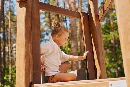 A child sits on a wooden play structure surrounded by trees. The sun shines brightly as the child looks out with curiosity, enjoying the moment