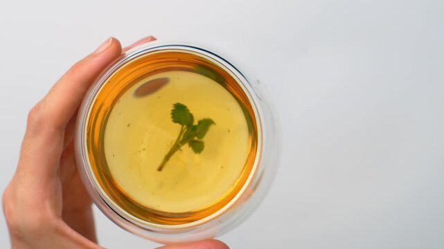 Top view of hand holding glass cup with organic mint drink. Single hand holding round double wall mug containing amber liquid and fresh green herb leaf on white surface.