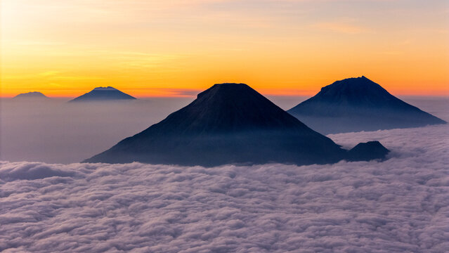 Aerial view of Gunung Bismo rising above a sea of clouds during a vibrant golden hour sunset in Kecamatan Kejajar, Jawa Tengah, Indonesia.