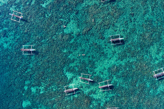 Aerial view of traditional outrigger boats floating in the clear turquoise waters over a coral reef with swimmers nearby in Kuta Selatan, Bali, Indonesia.