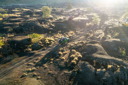 Aerial view of off-road jeeps driving through a rugged volcanic landscape with black lava fields and golden hour sunlight in Kuta Selatan, Bali, Indonesia.