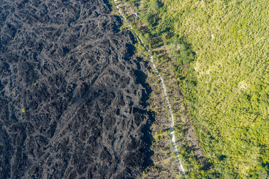 Aerial view of a dramatic landscape featuring a stark contrast between a black solidified lava flow and lush green forest separated by a narrow road in Kuta Selatan, Bali, Indonesia.