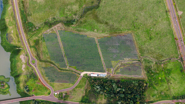 Aerial view of solar panels arranged in rows within a field, surrounded by green landscape, a river, and roads in Posadas, Misiones Province, Argentina.