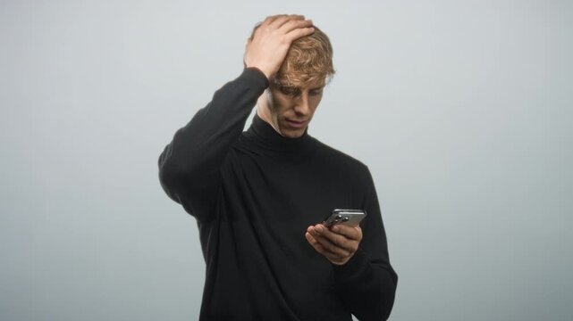 Young blond caucasian man in black turtleneck holding smartphone, looking down and touching head with hand in studio; confusion.