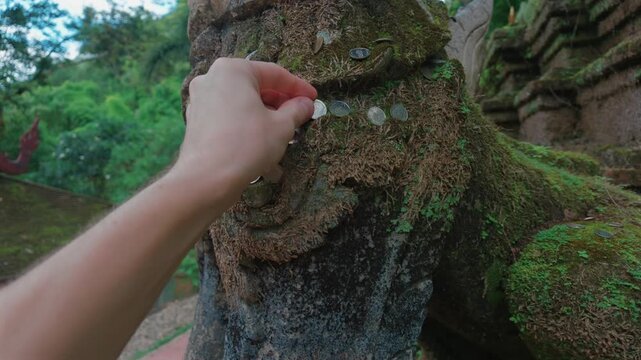 Hand placing coin on moss covered stone statue at Buddhist temple in Chiang Mai, Closeup of traditional coin offering ritual at sacred Thai shrine surrounded by nature 4k