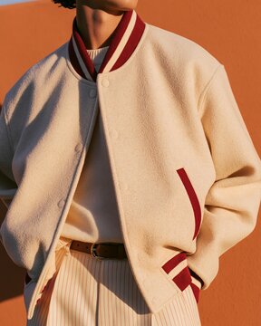 Close-up detail of cream wool varsity jacket with burgundy accents worn by a person against a warm orange background, showcasing stylish casual fashion elements