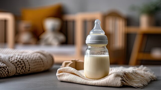 Close up of a glass baby bottle filled with milk on a cream colored napkin in a nursery with baby items in the background