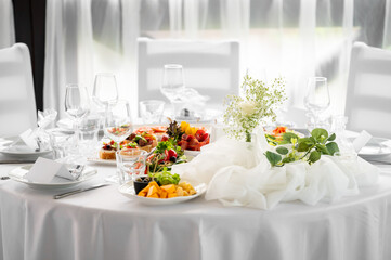 Fototapeta premium Elegant wedding table setting with white tablecloth, delicate flower centerpieces, wine glasses, and plates filled with gourmet appetizers and cheese platters in a brightly lit banquet hall.