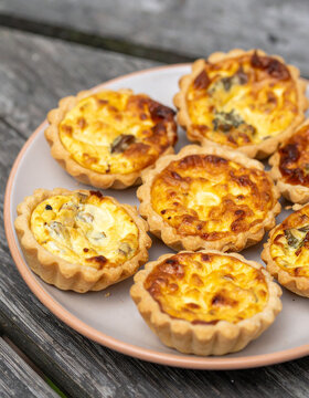 Freshly baked savory mini quiches on ceramic plate on rustic wooden picnic table, golden flaky crust, creamy filling and appetizing browned tops, delicious homemade meal, tasty snack.