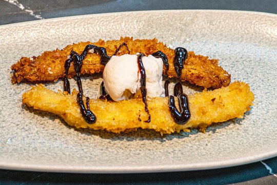  Professional chef hands plating a gourmet grilled banana dessert with vanilla ice cream and rich chocolate sauce.