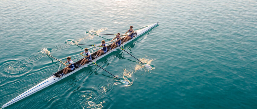 Rowing team practicing on calm lake water, aerial view of four athletes pulling oars in a competitive sweep boat under bright sunlight, ideal for sports editorial and team fitness concepts