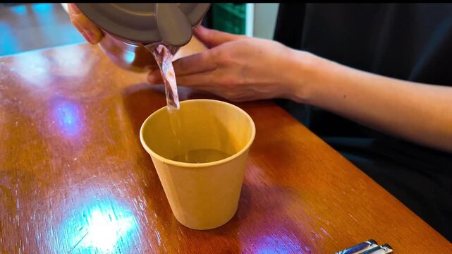 A girl pours water from an steel decanter into a disposable paper cup in a cafe.