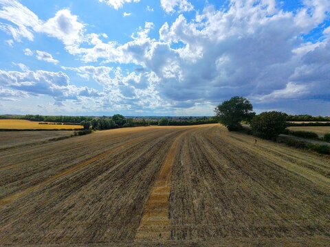 Aerial view of a harvested field with stubble, surrounded by hedgerows and trees under a dramatic cloudy sky in Burton Latimer, England, United Kingdom.
