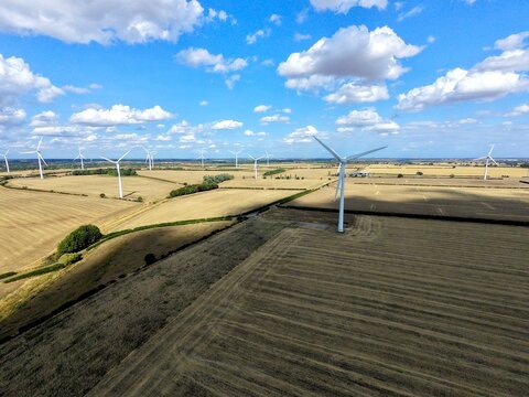 Aerial view of Burton Wold wind farm featuring multiple wind turbines standing in golden agricultural fields under a bright blue sky in Burton Latimer, England, United Kingdom.