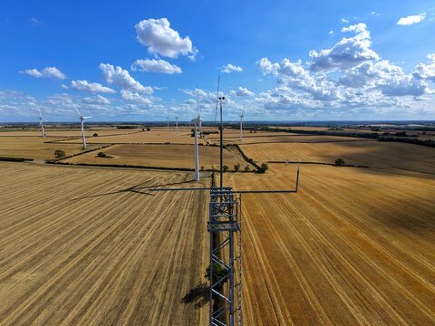 Aerial view of Burton Wold wind farm with a meteorological mast and wind turbines amidst golden wheat fields under a blue sky in Burton Latimer, England, United Kingdom.