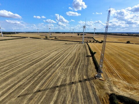 Aerial view of Burton Wold wind farm featuring turbine 1 and a tall meteorological mast in a harvested golden wheat field under a blue sky in Burton Latimer, England, United Kingdom.