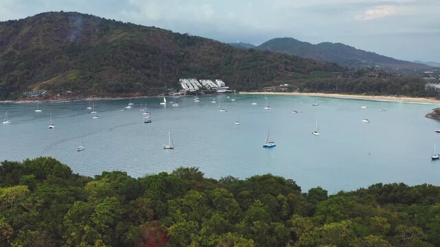 Sailboats anchored in calm bay of Nai Harn Beach, Phuket, Thailand, surrounded by lush green hills and sandy coastline under a cloudy sky. Tropical nature landscape. Aerial view drone flight panorama