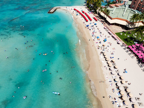 Aerial view of the turquoise waters and sandy beach of Oahu with people swimming and relaxing under red umbrellas at a resort in Honolulu County, Hawaii, United States.