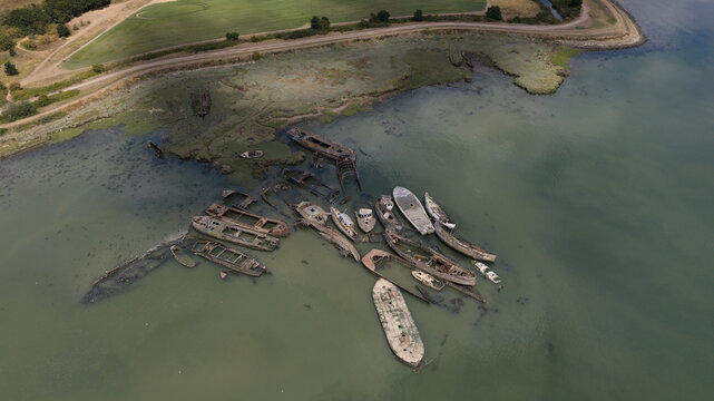 Aerial view of the Hoo Sailing Barge Graveyard with rusted wooden shipwrecks resting in the shallow tidal waters of the River Medway in Kent, United Kingdom.